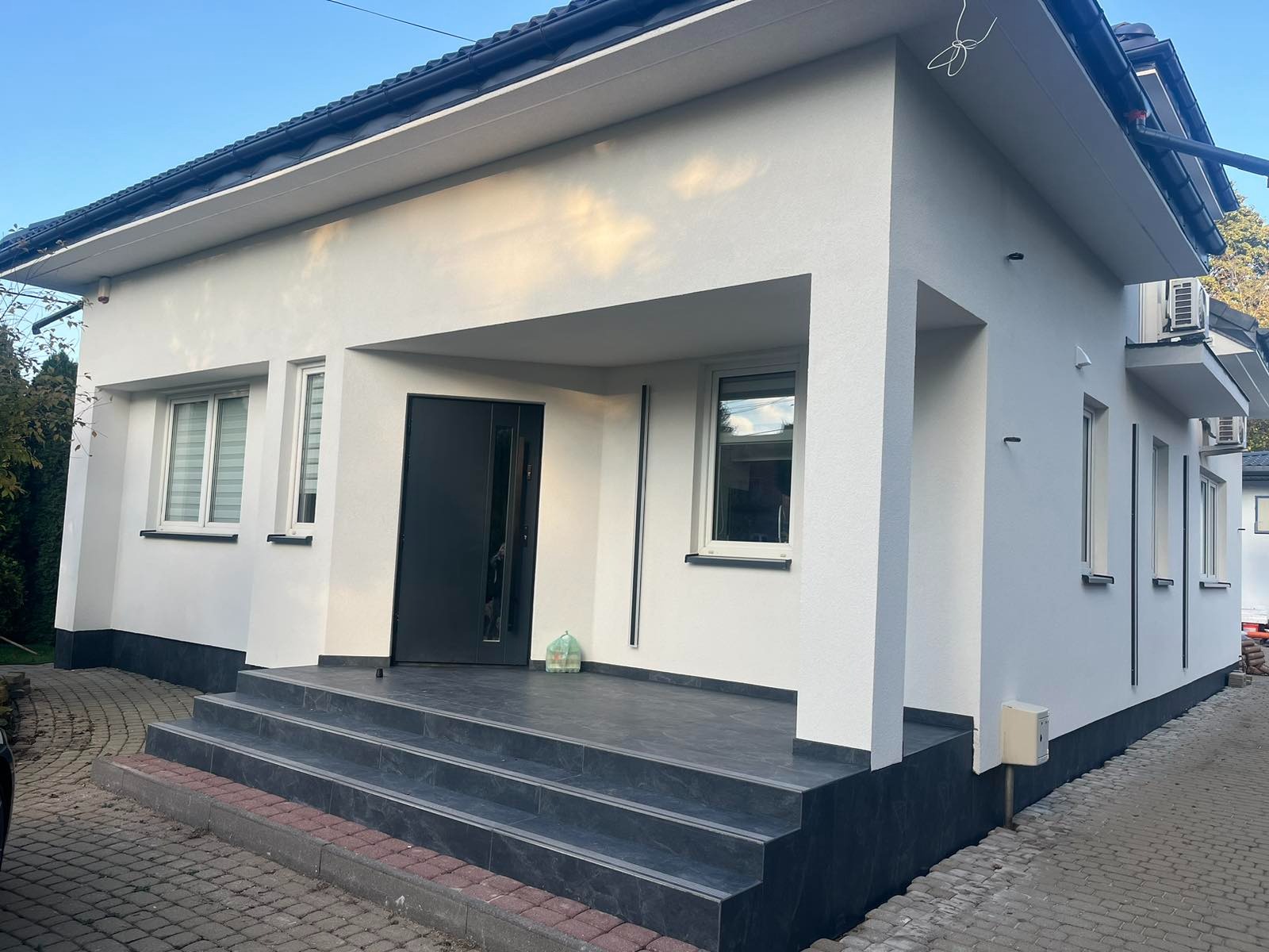 Wykończona elewacja domu jednorodzinnego z białym tynkiem strukturalnym, czarną stolarką okienną i drzwiami wejściowymi, oraz schodami wyłożonymi ciemnoszarymi płytkami imitującymi kamień.