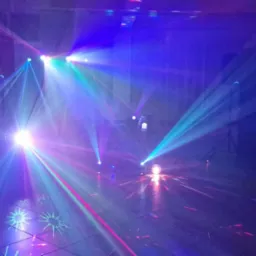 Sala oświetlona dynamicznymi, kolorowymi światłami punktowymi, tworzącymi wzory na podłodze, z widocznymi elementami konstrukcyjnymi sceny w tle.