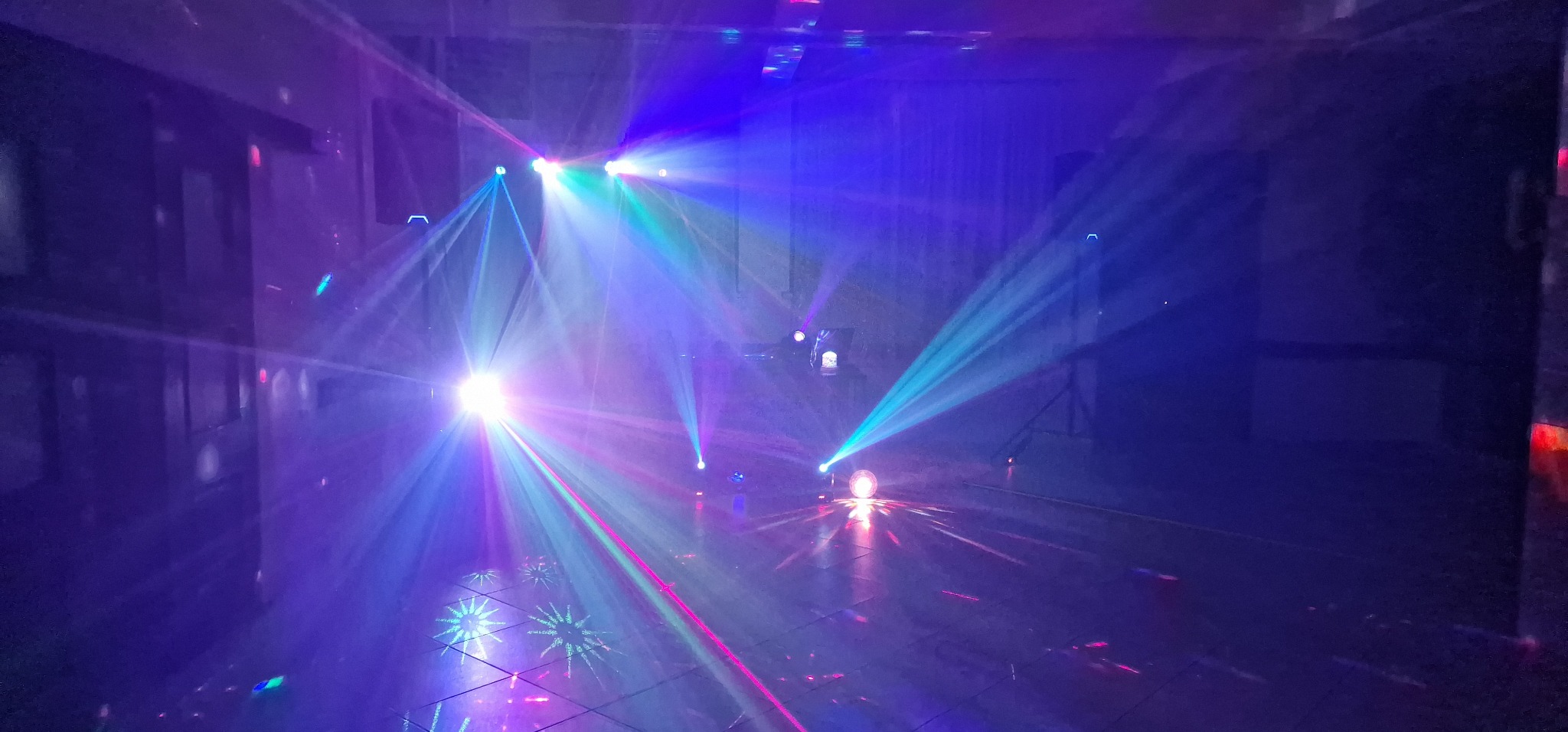 Sala oświetlona dynamicznymi, kolorowymi światłami punktowymi, tworzącymi wzory na podłodze, z widocznymi elementami konstrukcyjnymi sceny w tle.