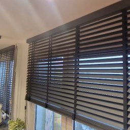 FTB SPÓŁKA Z OGRANICZONĄ ODPOWIEDZIALNOŚCIĄ - Ciemne żaluzje bambusowe 50mm zamontowane w oknach kuchni, widoczne z bliska, z elementami wyposażenia wnętrza w tle. Ustawione poziomo, częściowo zasłonięte.