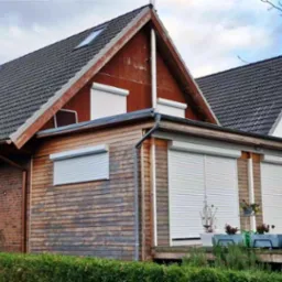 Dom jednorodzinny z drewnianą elewacją i białymi roletami zewnętrznymi opuszczonymi na oknach, widoczne rośliny w donicach na tarasie.