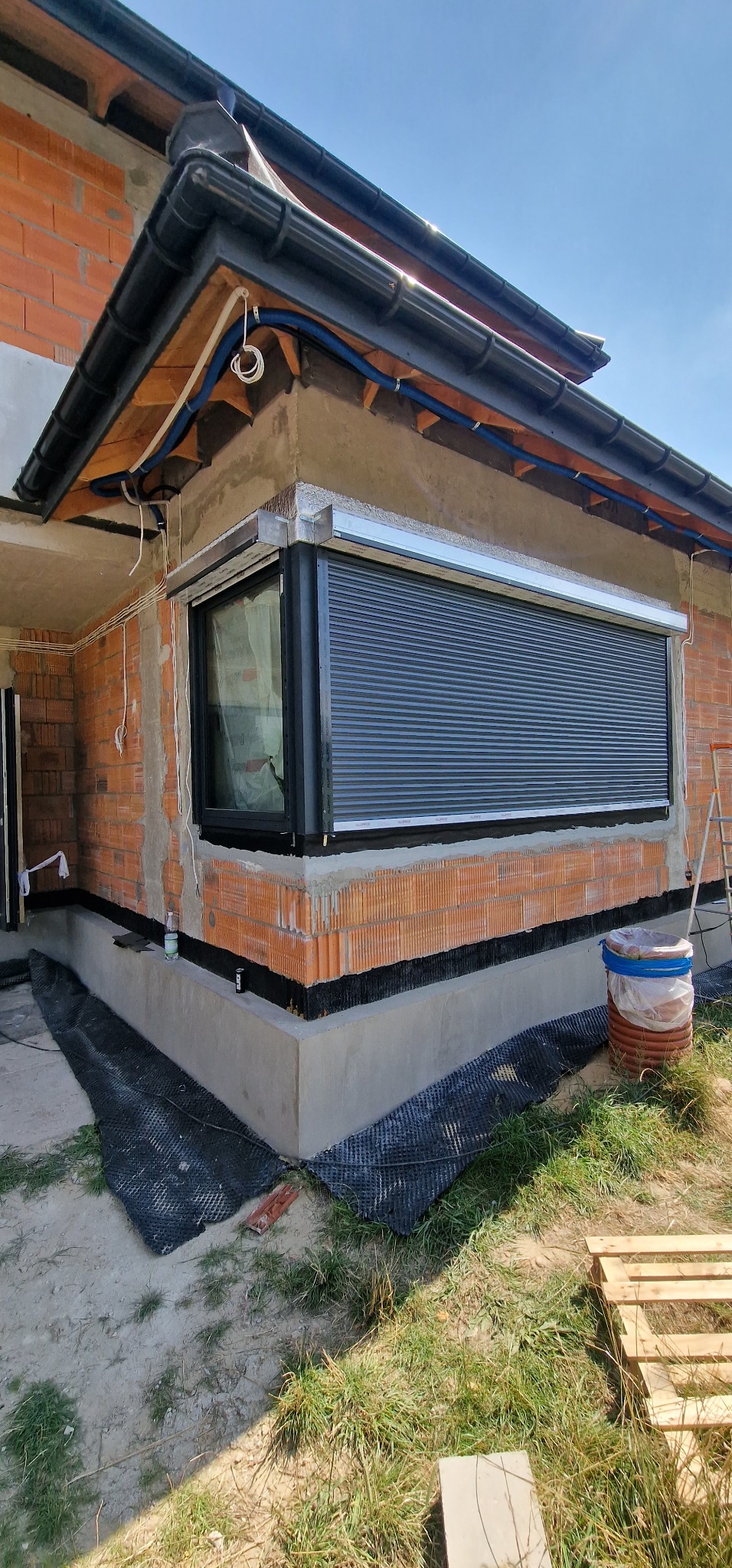 Ciemna roleta zewnętrzna zamontowana na oknie w surowym budynku z czerwonej cegły. Widoczne elementy instalacji i fragment drabiny.