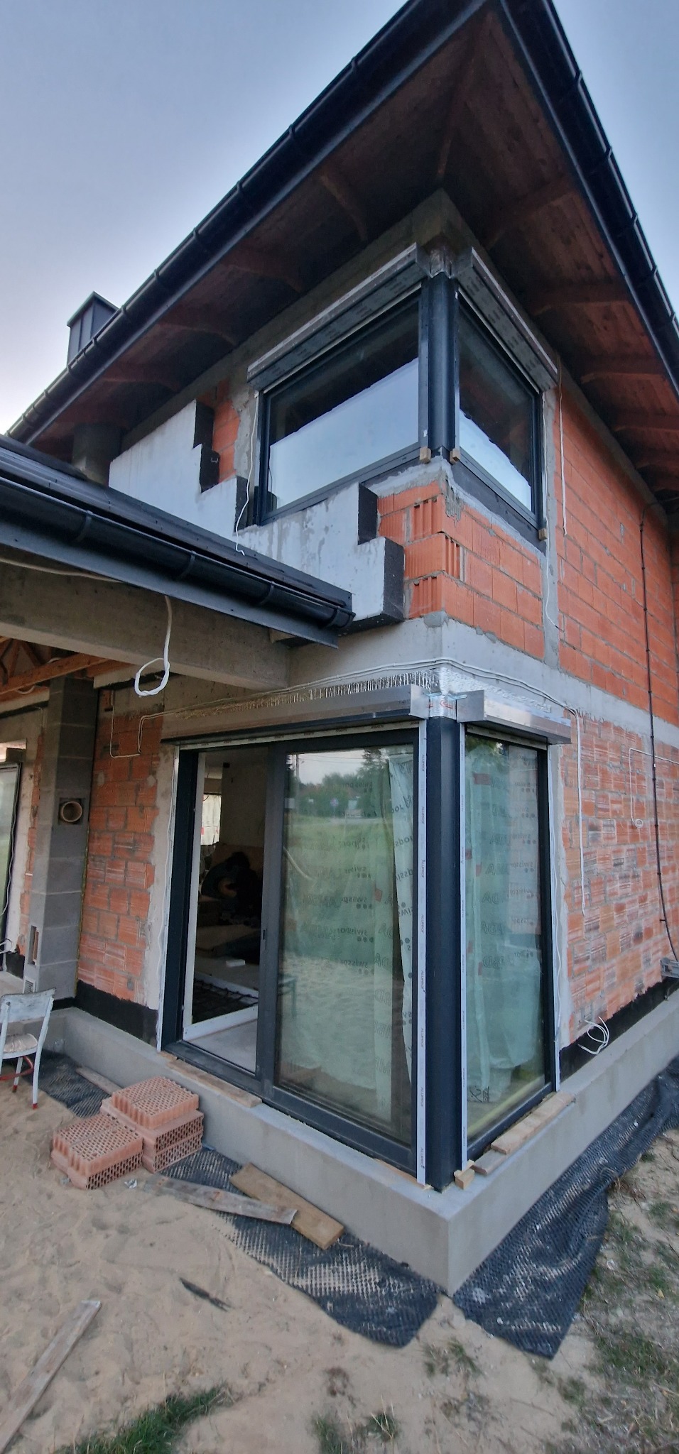 Dom w trakcie budowy z zamontowanymi oknami PCV z roletami zewnętrznymi. Widoczne niedokończone elewacje i materiały budowlane.