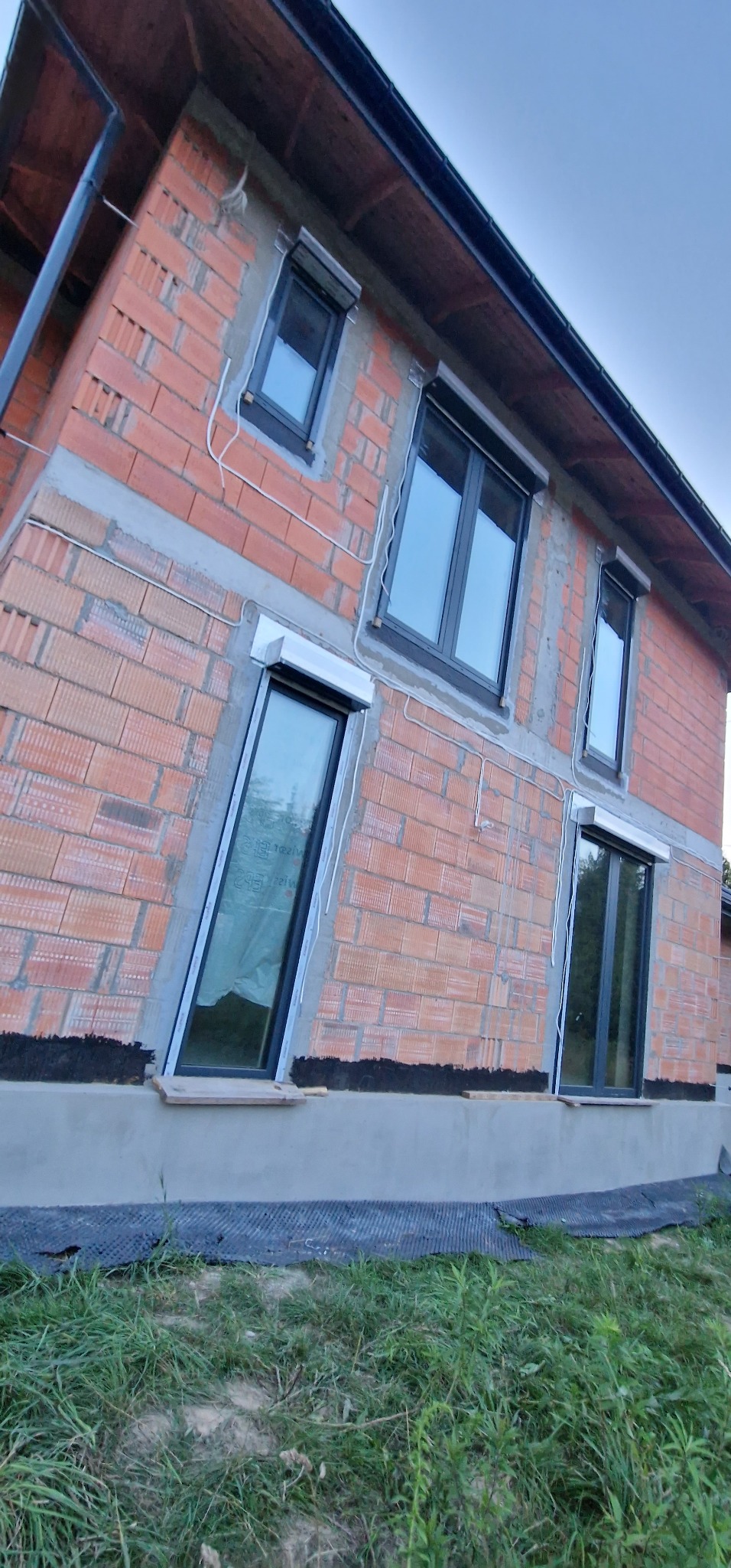 Surowy budynek z czarnymi oknami i częściowo opuszczonymi roletami zewnętrznymi. Widoczne przewody elektryczne na elewacji. Ujęcie z dołu.