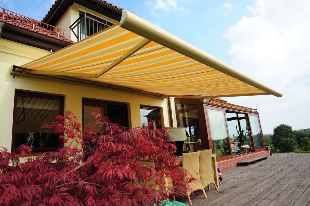 Żółto-biała markiza tarasowa z wysuwanym ramieniem, zamontowana nad oknami i przeszklonym tarasem domu. Czerwony klon palmowy w pierwszym planie.