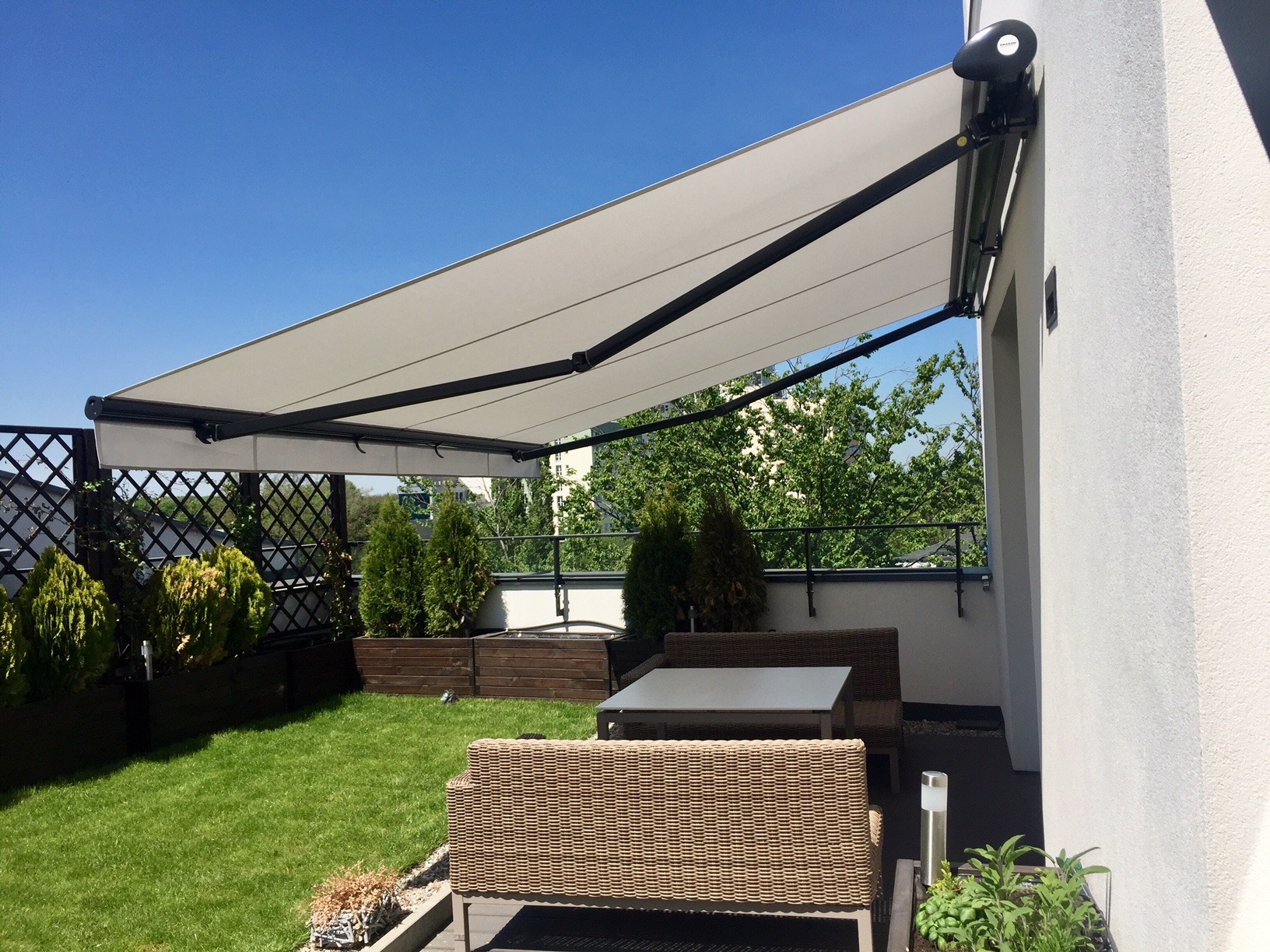 Biała markiza tarasowa z czarną konstrukcją na tle błękitnego nieba, widok z ogrodu na balkonie z meblami ogrodowymi i zielenią.