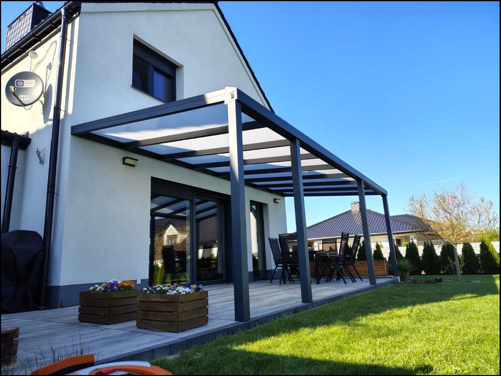 Nowoczesna, szara pergola tarasowa z przezroczystym zadaszeniem, przylegająca do białego domu z dużymi oknami. Widoczne drewniane donice z kwiatami i zadbany trawnik.