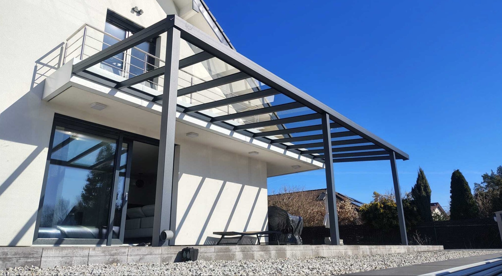 Nowoczesna, szara pergola tarasowa z przezroczystym zadaszeniem, przylegająca do białego domu z dużym oknem przesuwnym. Błękitne niebo w tle.