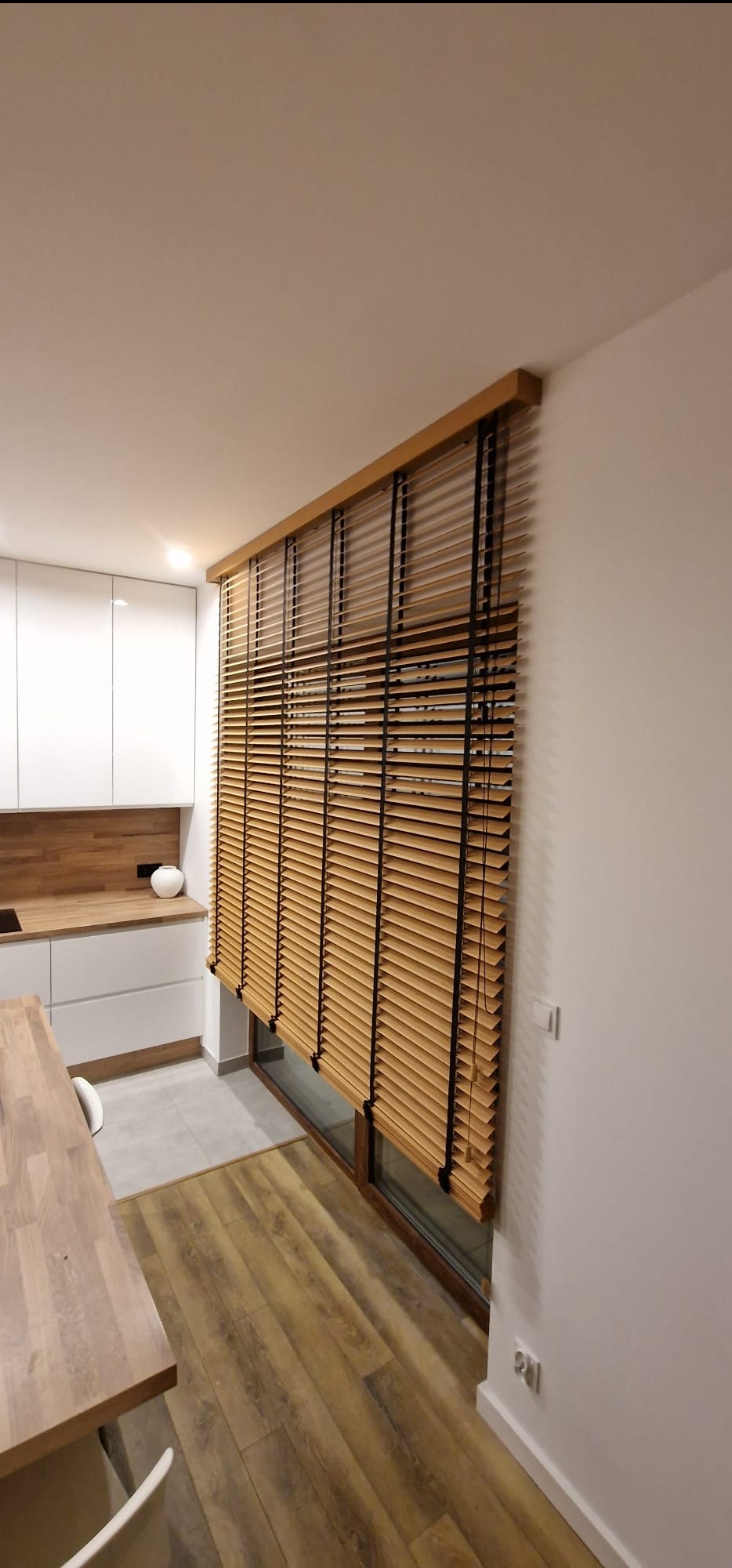 Bambusowe żaluzje poziome w jasnym wnętrzu kuchni, zamontowane na oknie. Naturalne drewno, minimalistyczny design, widok z perspektywy pomieszczenia.