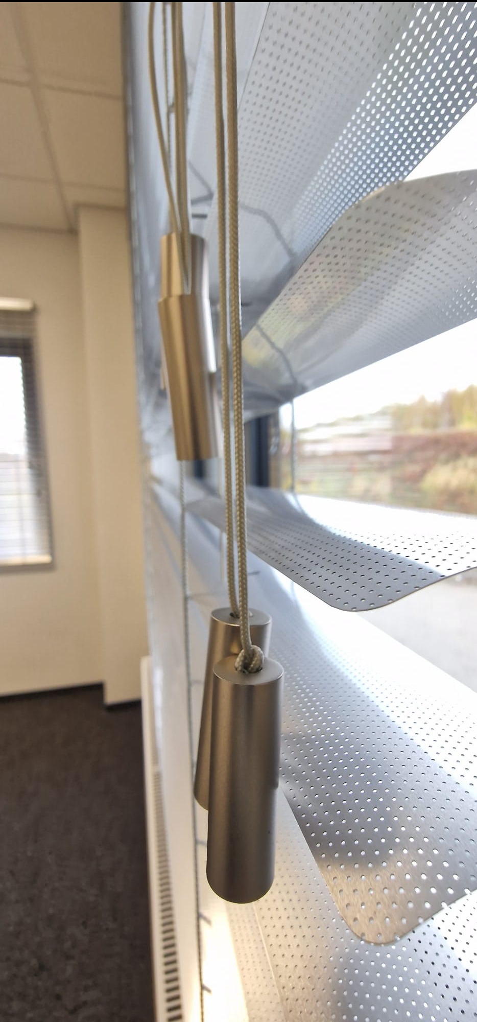 Metalowe żaluzje perforowane z widocznymi sznurkami i obciążnikami, zamontowane w oknie. Widok z bliska, z zewnątrz widoczne drzewa.