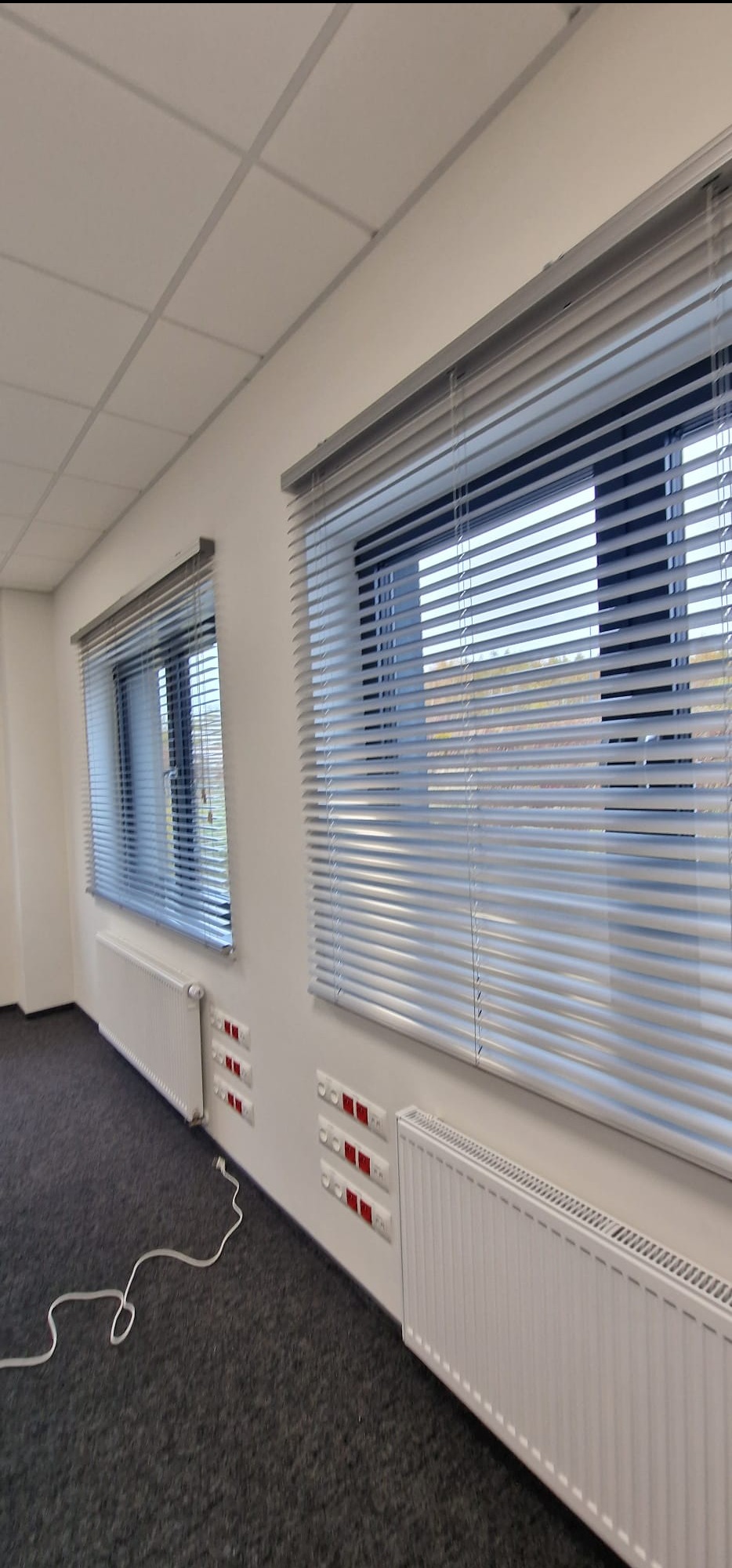 Dwa okna z aluminiowymi żaluzjami poziomymi w biurze. Widoczne grzejniki pod oknami i szary dywan. Minimalistyczny wystrój wnętrza, jasne ściany i sufit.