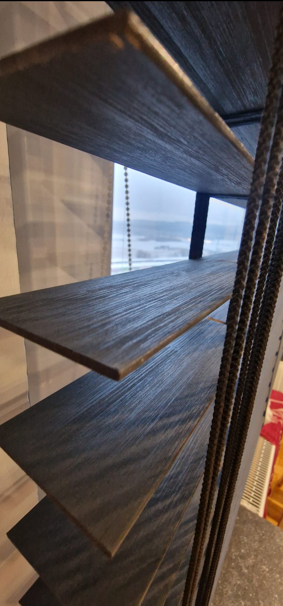 Ciemne żaluzje bambusowe 50mm z widokiem na zimowy krajobraz za oknem. Detal sznurków sterujących i faktury drewna. Ujęcie z dołu, podkreślające głębię perspektywy.