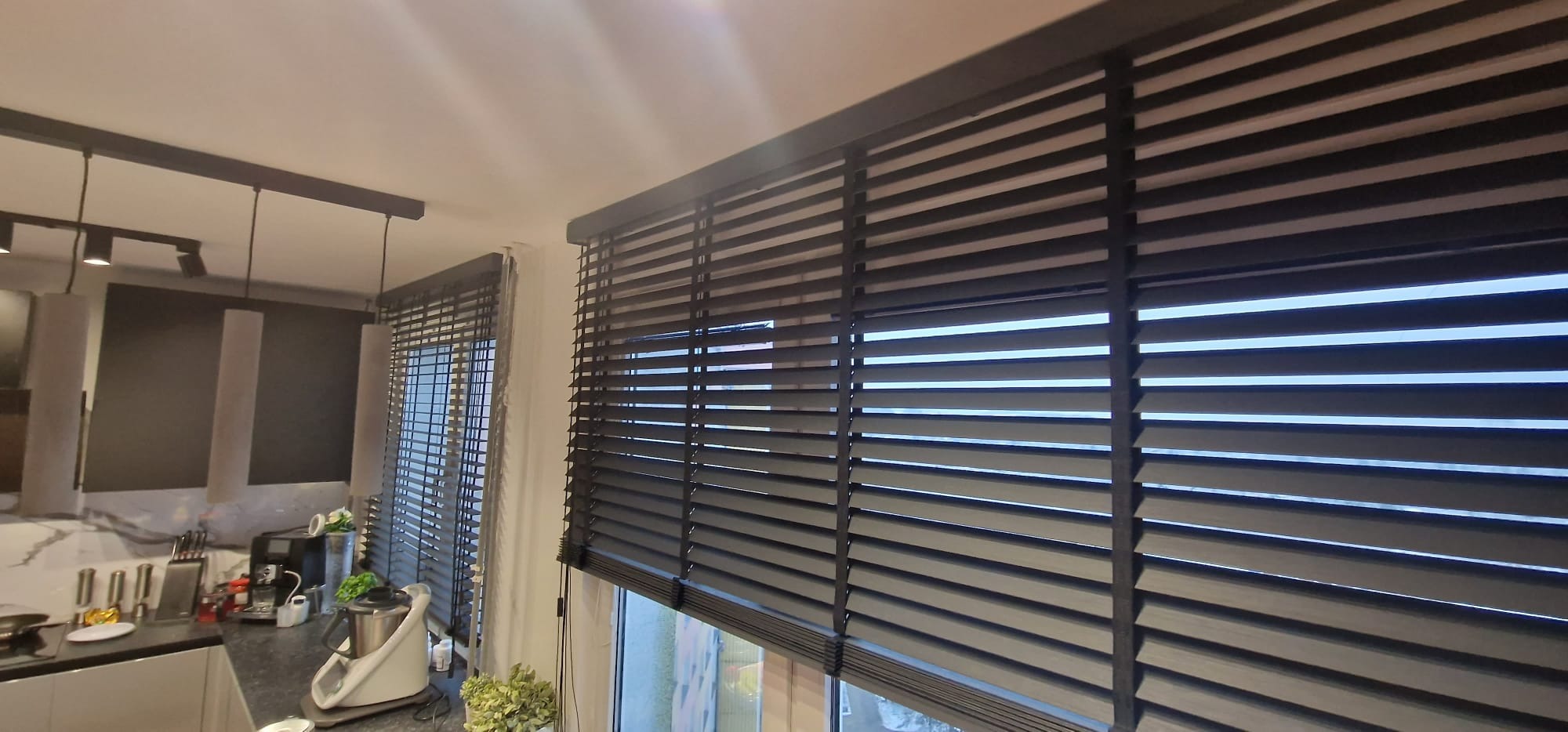 Ciemne żaluzje bambusowe 50mm zamontowane w oknach kuchni, widoczne z bliska, z elementami wyposażenia wnętrza w tle. Ustawione poziomo, częściowo zasłonięte.