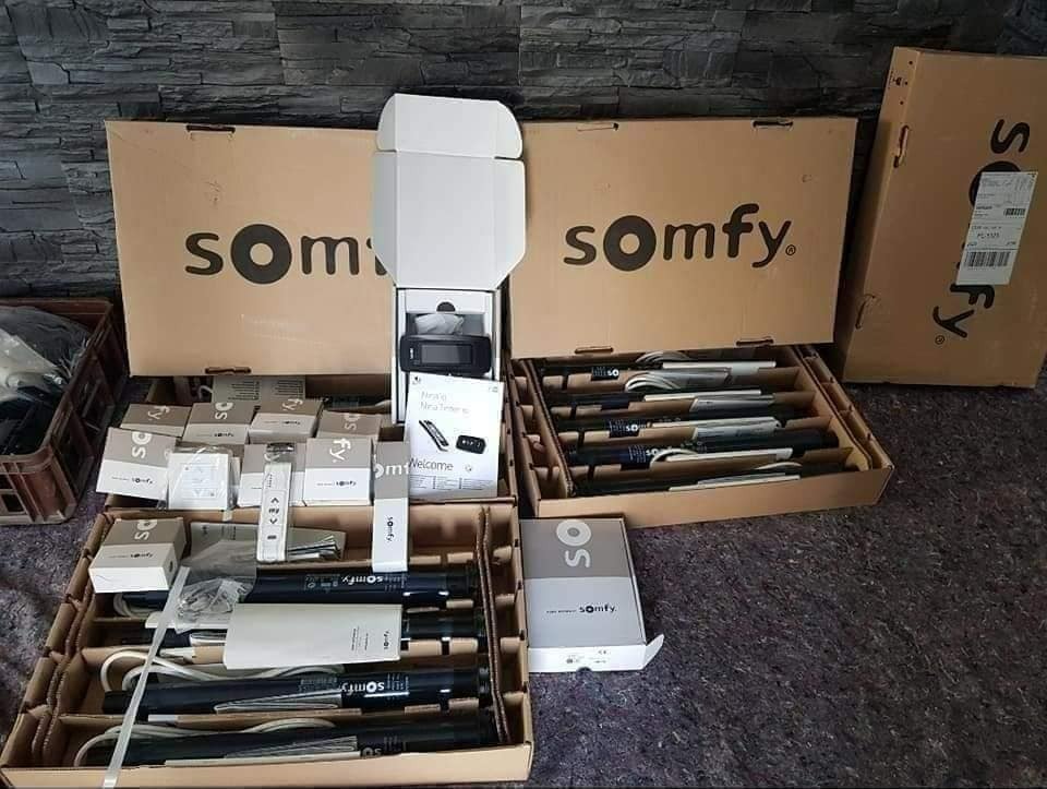 Rozpakowane komponenty systemu sterowania roletami marki Somfy, widoczne silniki, piloty, centralka i opakowania kartonowe z logo firmy na tle szarej ściany.