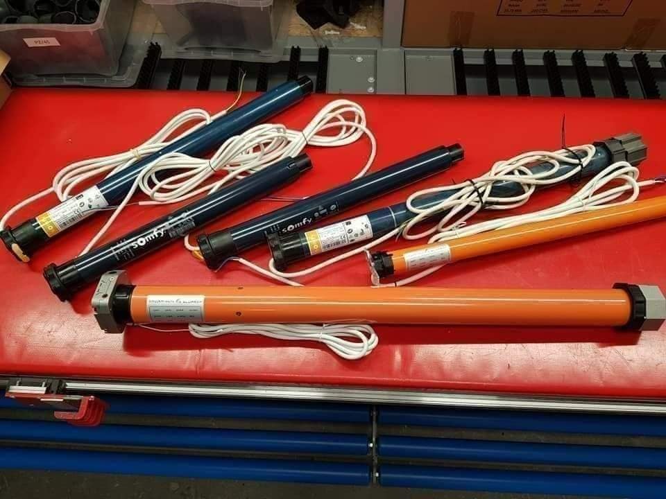 Silniki rurowe Somfy do rolet antywłamaniowych i zewnętrznych, niebieskie i pomarańczowe, leżą na czerwonym warsztatowym stole.