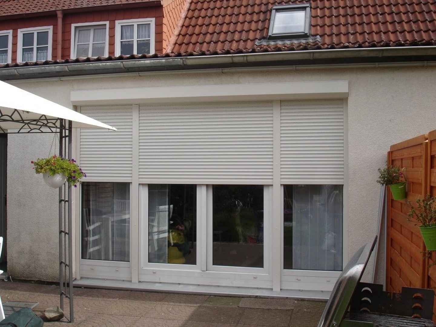 Białe rolety zewnętrzne opuszczone na oknach w domu jednorodzinnym, widoczne doniczki z kwiatami na zewnątrz.