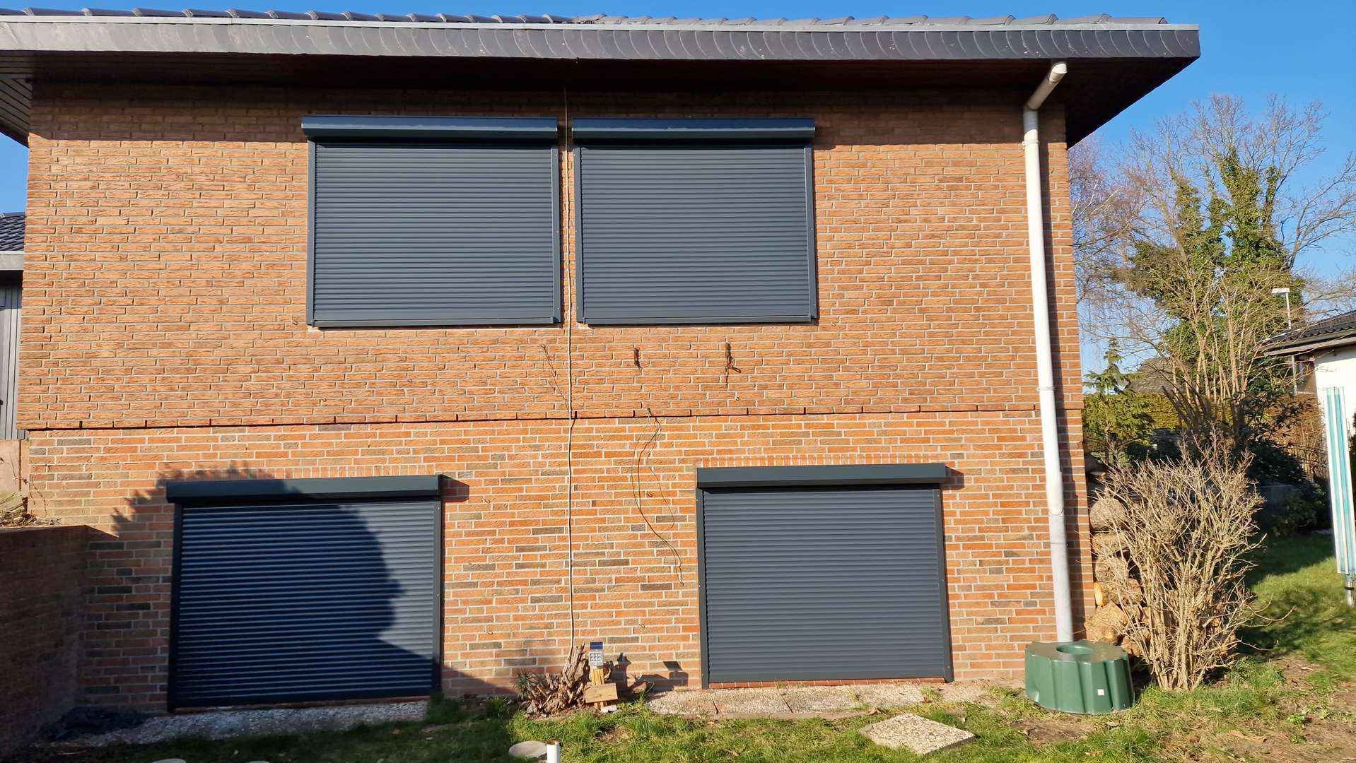 Ceglana elewacja domu z trzema szarymi roletami zewnętrznymi, dwie na górze i jedna na dole, z widocznymi przewodami elektrycznymi na ścianie i fragmentem ogrodu.