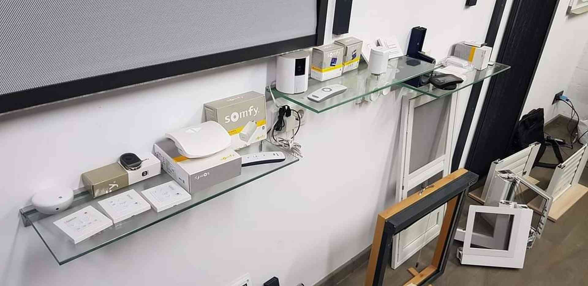 Ekspozycja produktów Somfy na szklanych półkach w salonie w Wałbrzychu, wraz z ramami okiennymi i elementami drzwiowymi w dolnej części kadru.