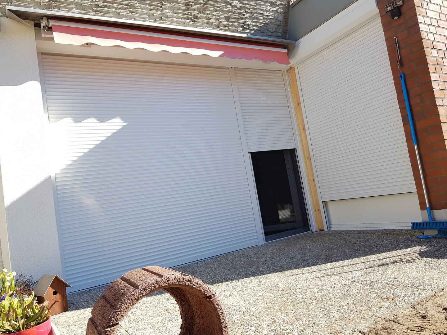 Białe rolety zewnętrzne z częściowo odsłoniętym oknem, widoczne narzędzia i dekoracyjny element ogrodowy na pierwszym planie.