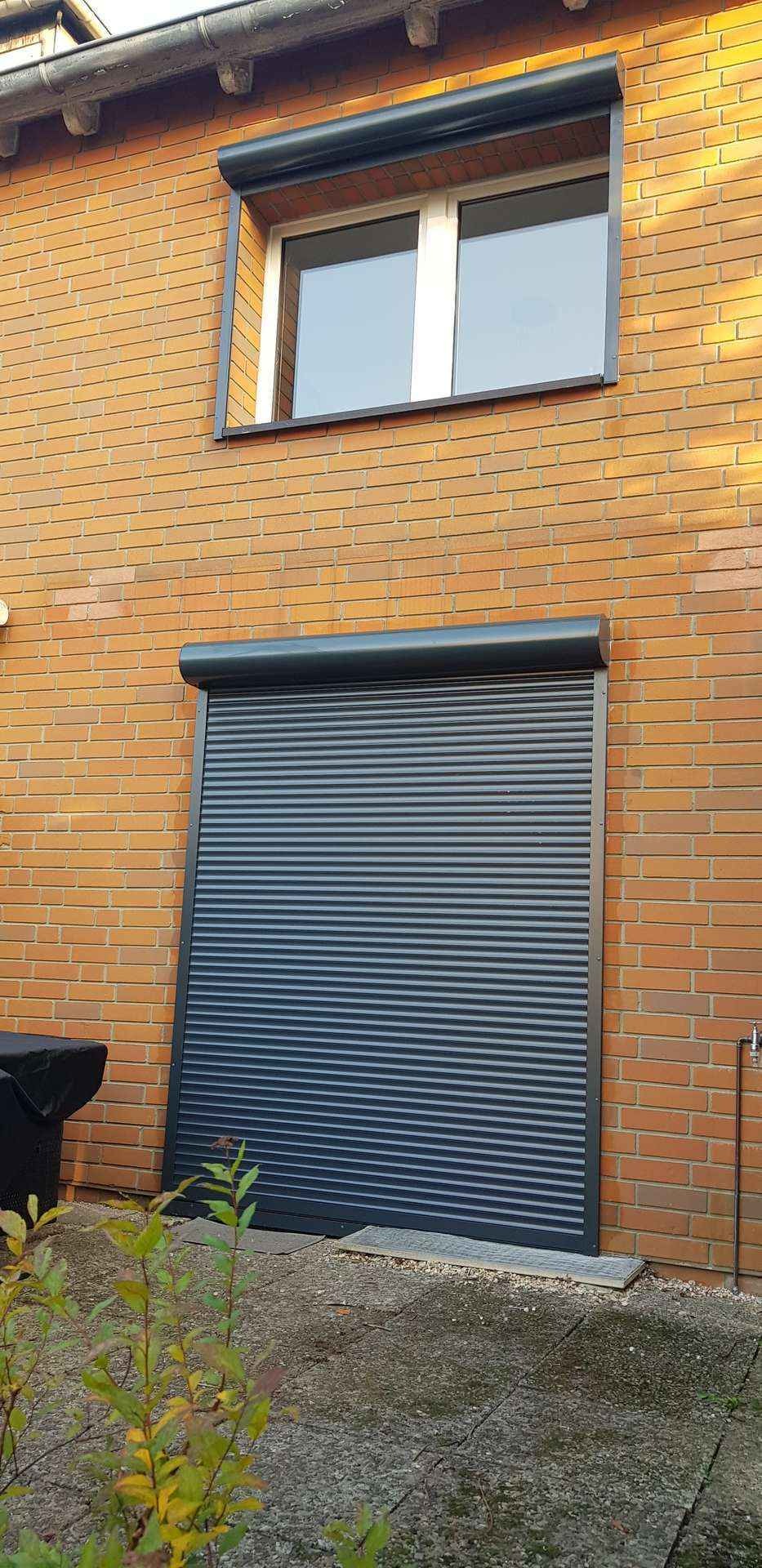 Ciemnoszara roleta zewnętrzna opuszczona na okno na tle ceglanej ściany budynku, widoczny fragment okna powyżej z podobną roletą.