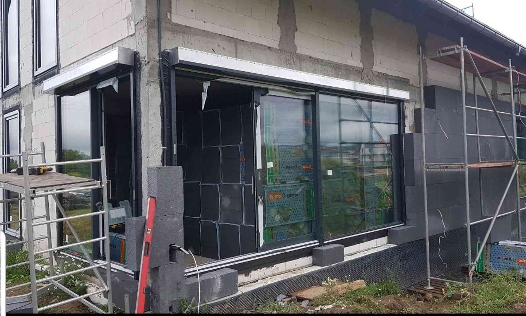 Montaż okna przesuwnego z roletą zewnętrzną w trakcie ocieplania budynku, widoczne rusztowanie, poziomica i materiały izolacyjne.