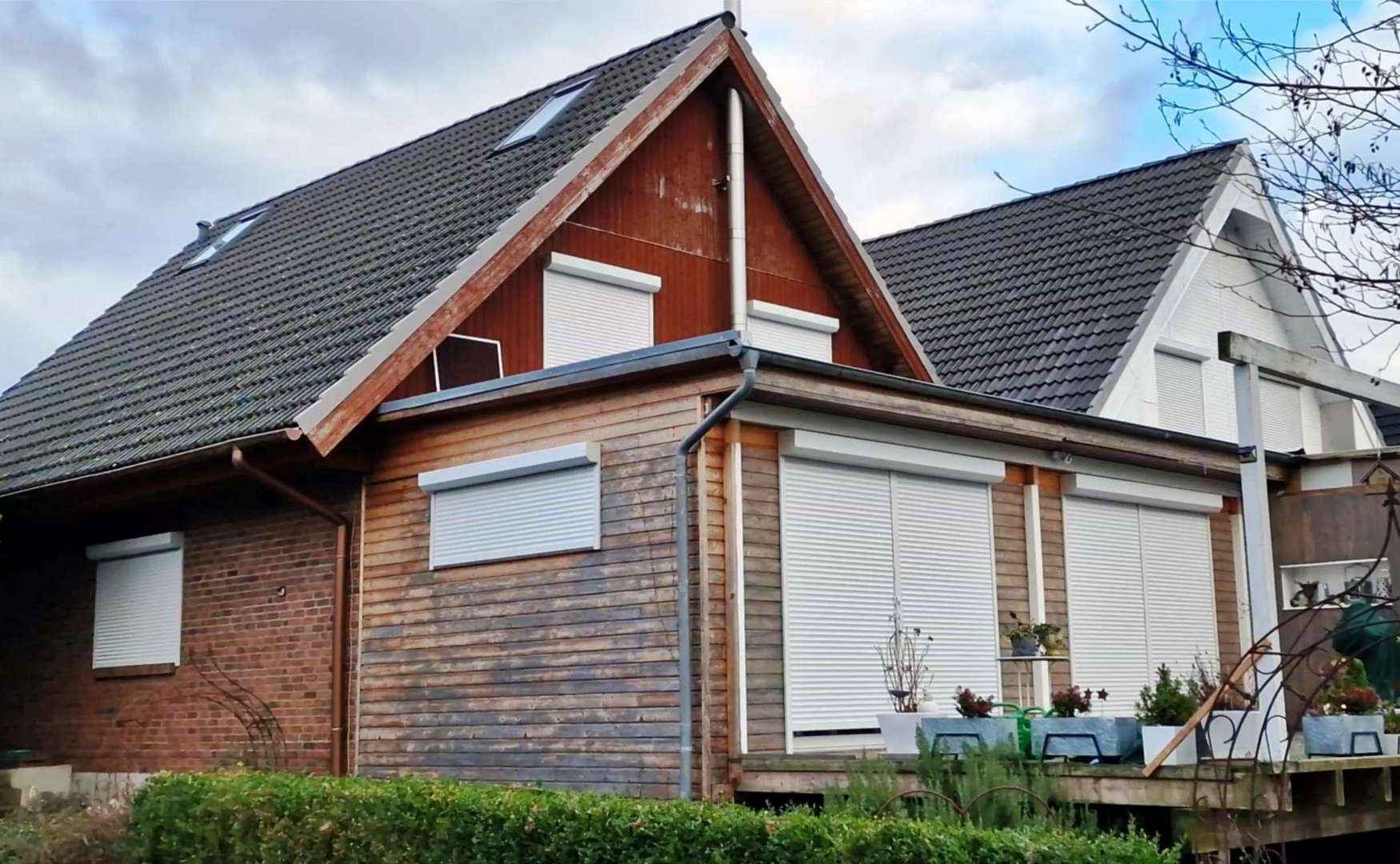 Dom jednorodzinny z drewnianą elewacją i białymi roletami zewnętrznymi opuszczonymi na oknach, widoczne rośliny w donicach na tarasie.