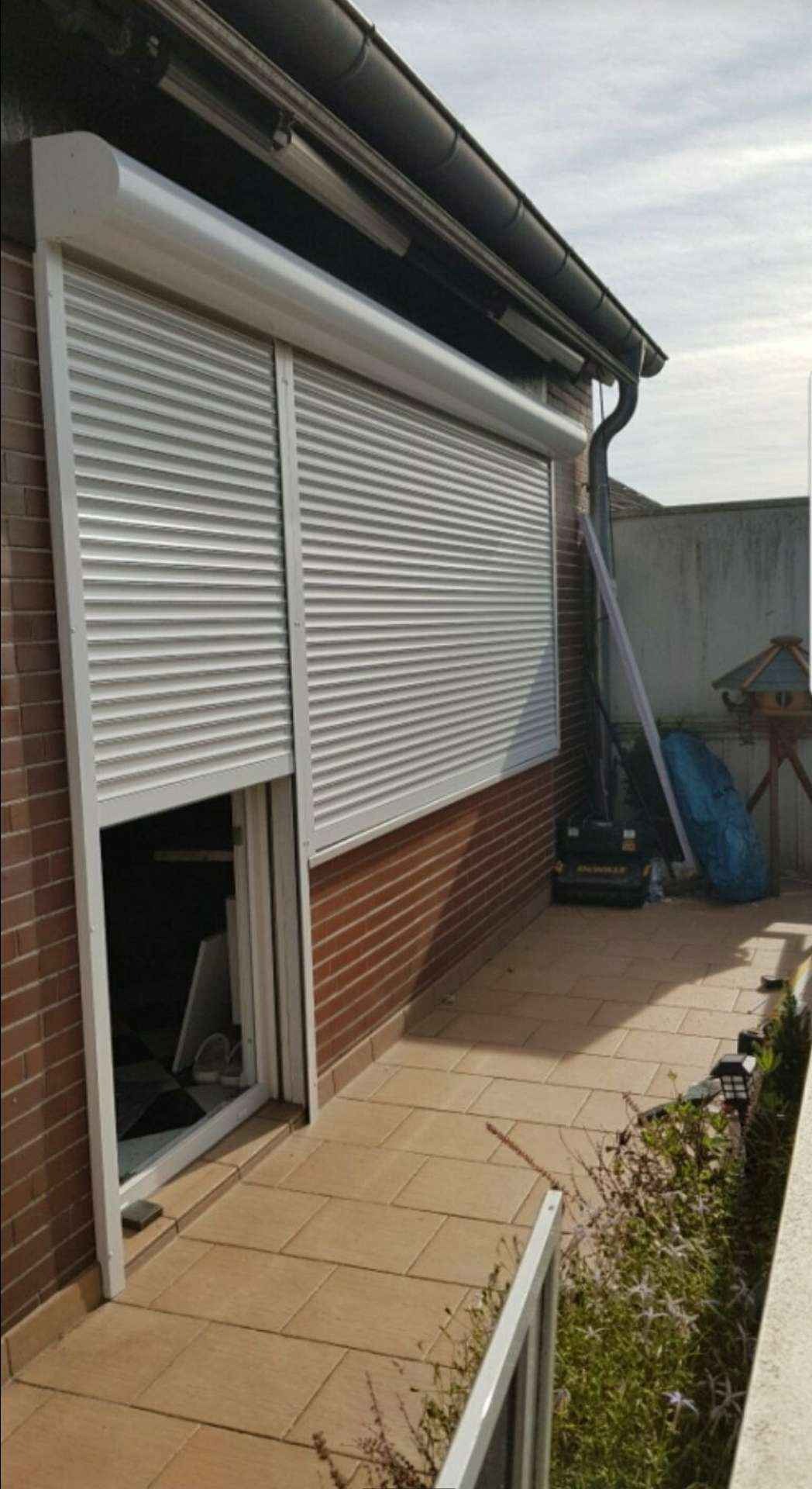 Częściowo opuszczona, biała roleta zewnętrzna na oknie z ceglaną ścianą, widoczny fragment podwórka z kostką brukową i sprzętem budowlanym.