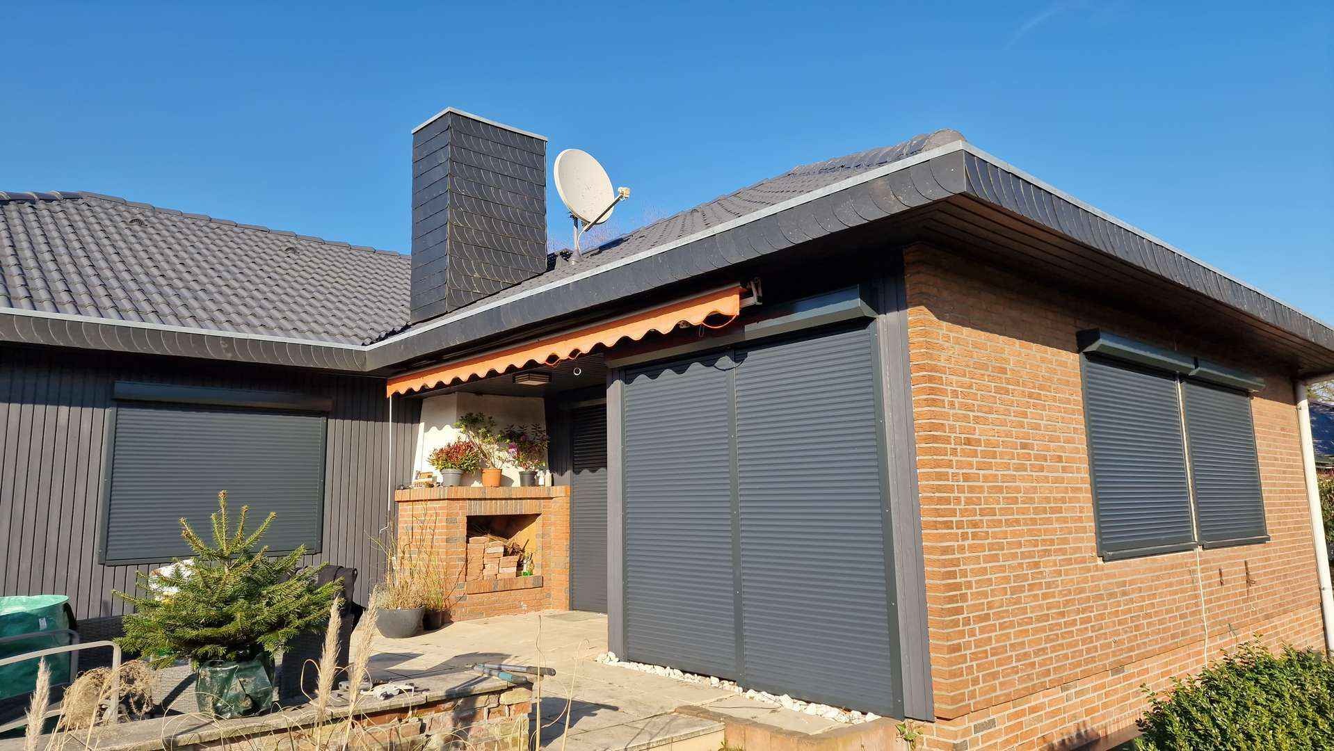 Dom jednorodzinny z szarymi roletami zewnętrznymi na oknach i drzwiach tarasowych, widoczne również ceglana elewacja i komin z anteną satelitarną.