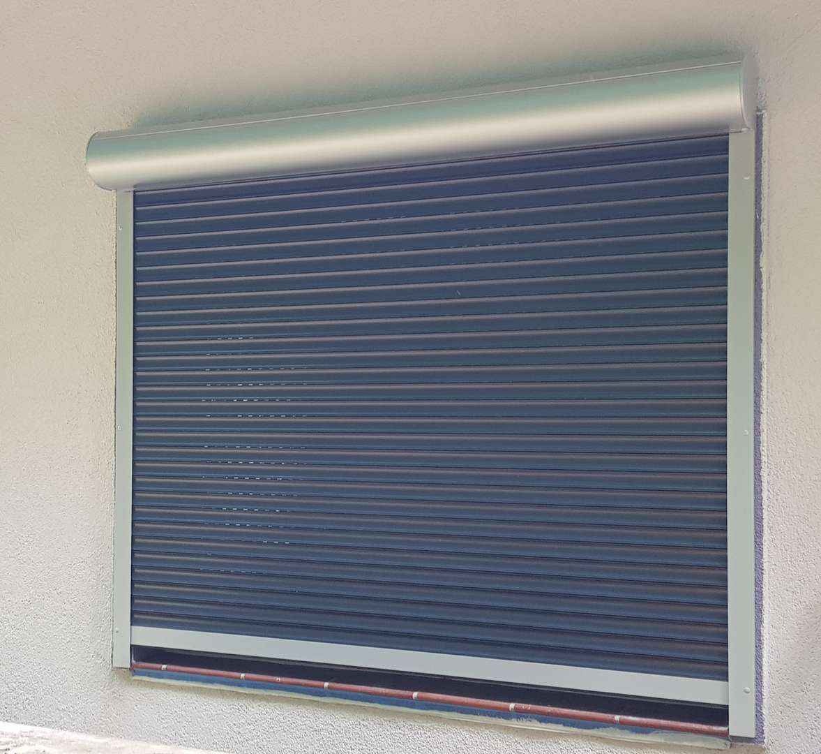 Zasunięta, ciemna roleta zewnętrzna z aluminiową obudową na tle jasnego, chropowatego muru.