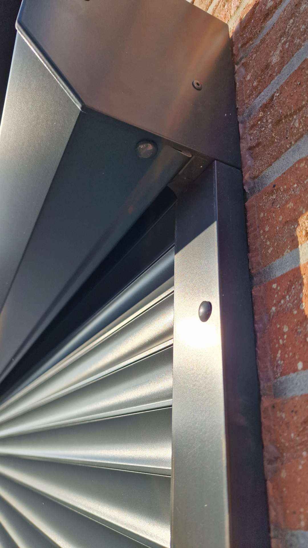 Fragment zamkniętej, grafitowej rolety zewnętrznej z widocznym mocowaniem do ściany z czerwonej cegły, uchwycony z bliskiej perspektywy.