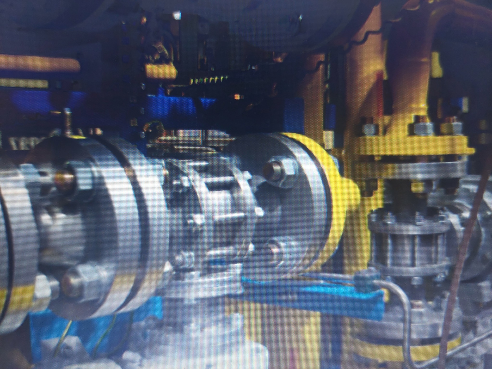 Fragment instalacji przemysłowej z rurami i zaworami, z żółtymi i niebieskimi elementami podtrzymującymi, widoczne śruby i połączenia kołnierzowe.