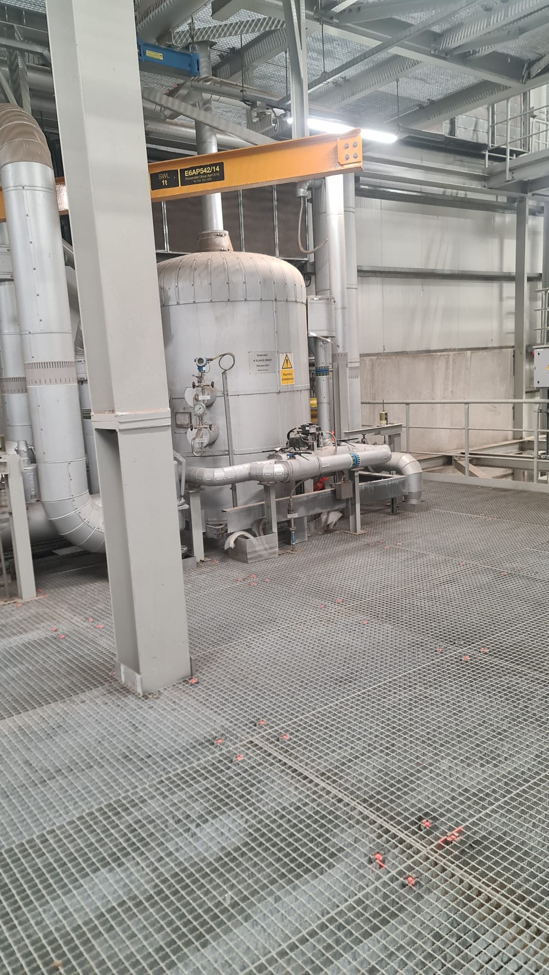 Widok na przemysłowy zbiornik ciśnieniowy z rurami i zaworami, umieszczony na metalowej platformie kratowej w hali produkcyjnej, z widocznymi elementami konstrukcji stalowej i instalacji sufitowych.