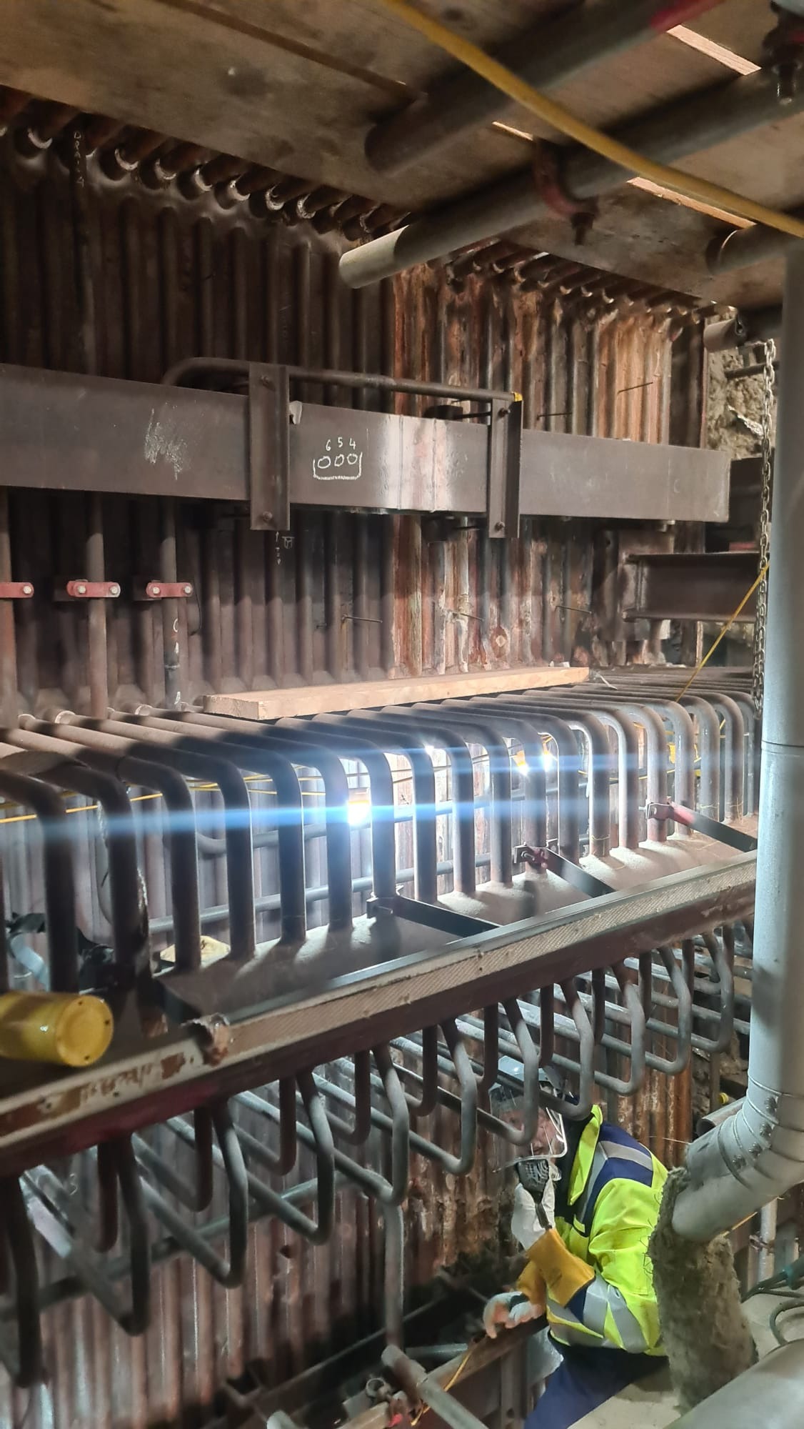 Prace spawalnicze wewnątrz kotła przemysłowego, widoczne rury, metalowa konstrukcja z napisem '654 1000', spawacz w kombinezonie ochronnym i przyłbicy, iskry spawania.