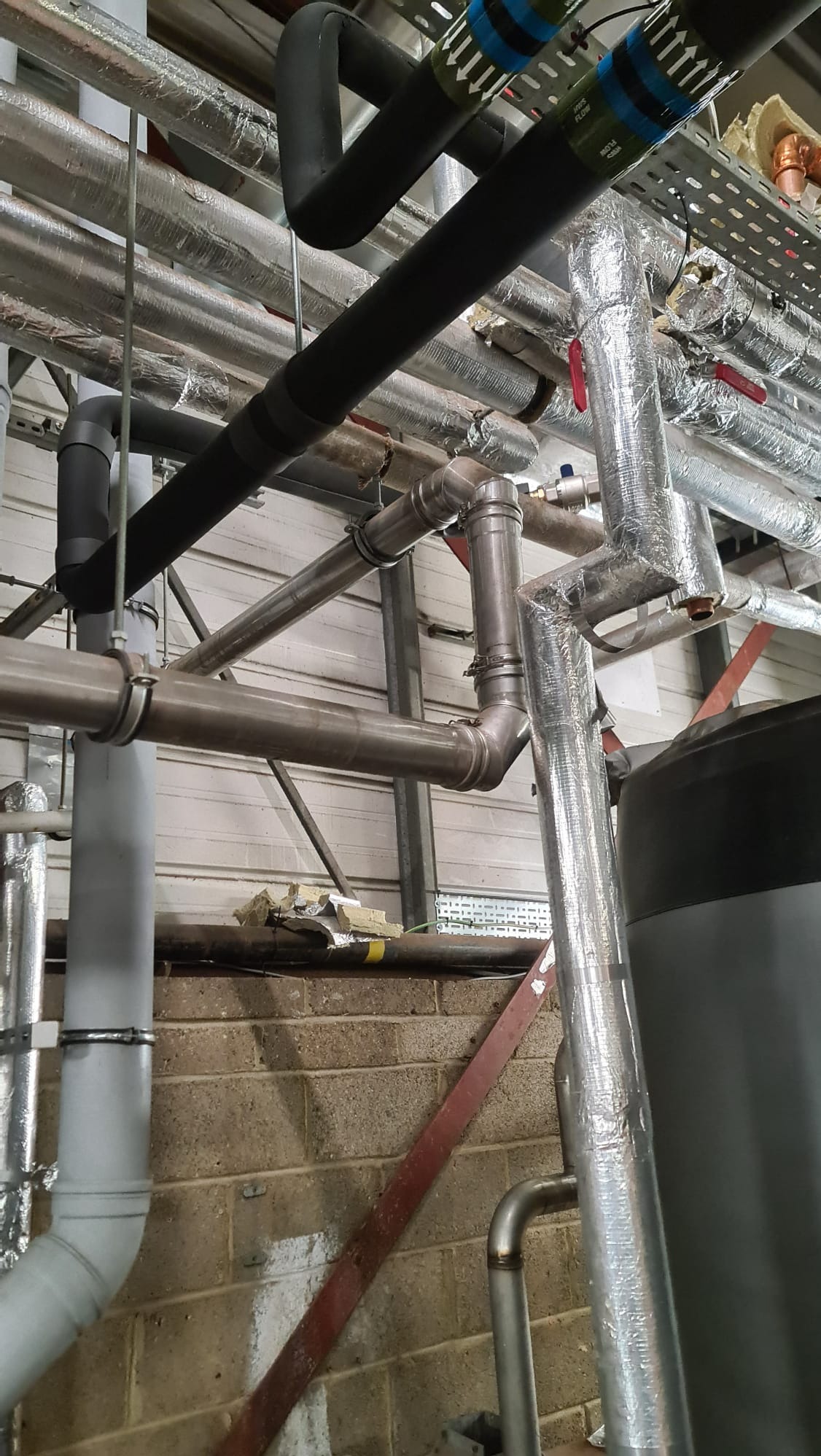 Złożony system rur instalacji przemysłowej z izolacją termiczną, obejmujący rury stalowe, miedziane i z tworzywa sztucznego, widoczne zawory i elementy mocujące, na tle surowej ściany z cegły...