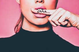 Kobieta z palcem wskazującym na ustach, na palcu napis 'Shhh...', różowe tło.