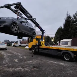 Szara osobówka podnoszona przez ramię żółtej lawety na tle zabudowań i drzew, widoczny numer rejestracyjny pojazdu oraz napisy na lawecie: Abschleppdienst, Unfallservice, Mietwagen.