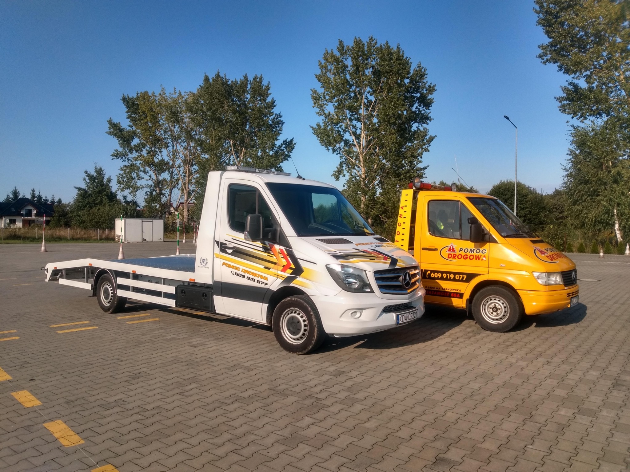 Dwa samochody lawety: biały Mercedes z czarno-żółtymi akcentami i żółty Mercedes z napisem 'Pomoc Drogowa', zaparkowane w Dąbrowie Tarnowskiej na kostce brukowej w słoneczny dzień.