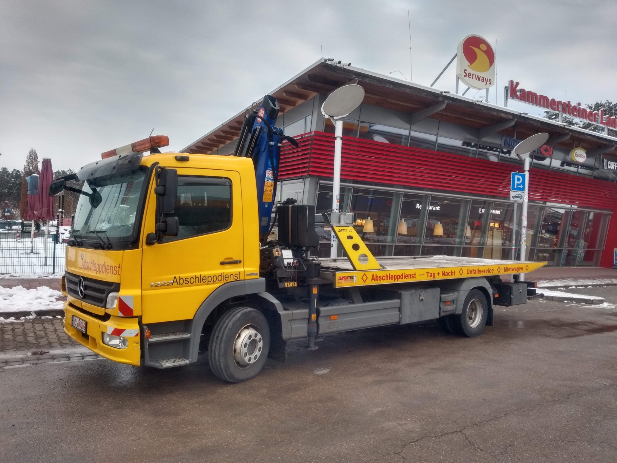 Żółta ciężarówka pomocy drogowej Mercedes-Benz z niebieskim dźwigiem, zaparkowana na parkingu przed budynkiem restauracji Serways Kammersteiner Land. Na karoserii widoczne napisy 'Abschleppdienst'...
