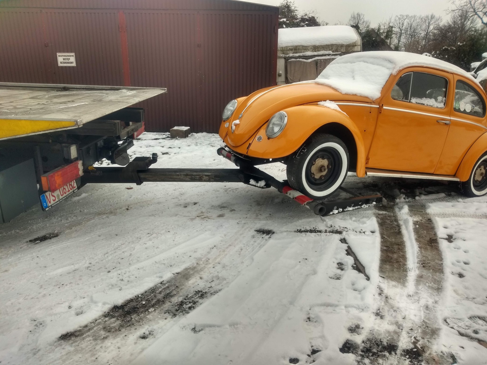Pomarańczowy Volkswagen Garbus z białymi oponami, częściowo pokryty śniegiem, załadowany na lawetę w zimowej scenerii. W tle widoczny brązowy blaszany garaż i tabliczka z napisem 'Wstęp wzbroniony'.