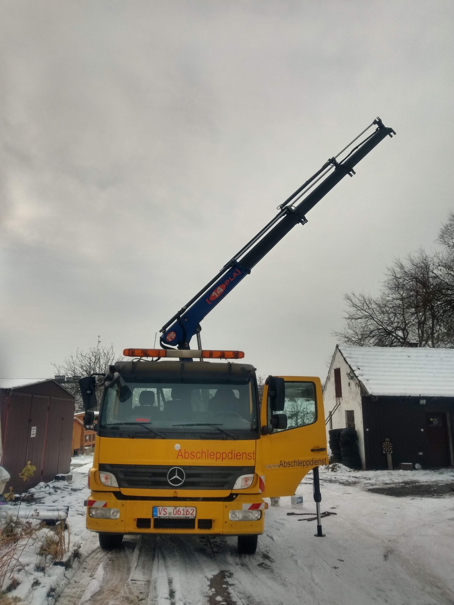 Żółty pojazd pomocy drogowej z wysięgnikiem na tle zaśnieżonego terenu i zabudowań gospodarczych, otwarte drzwi z widocznym stabilizatorem