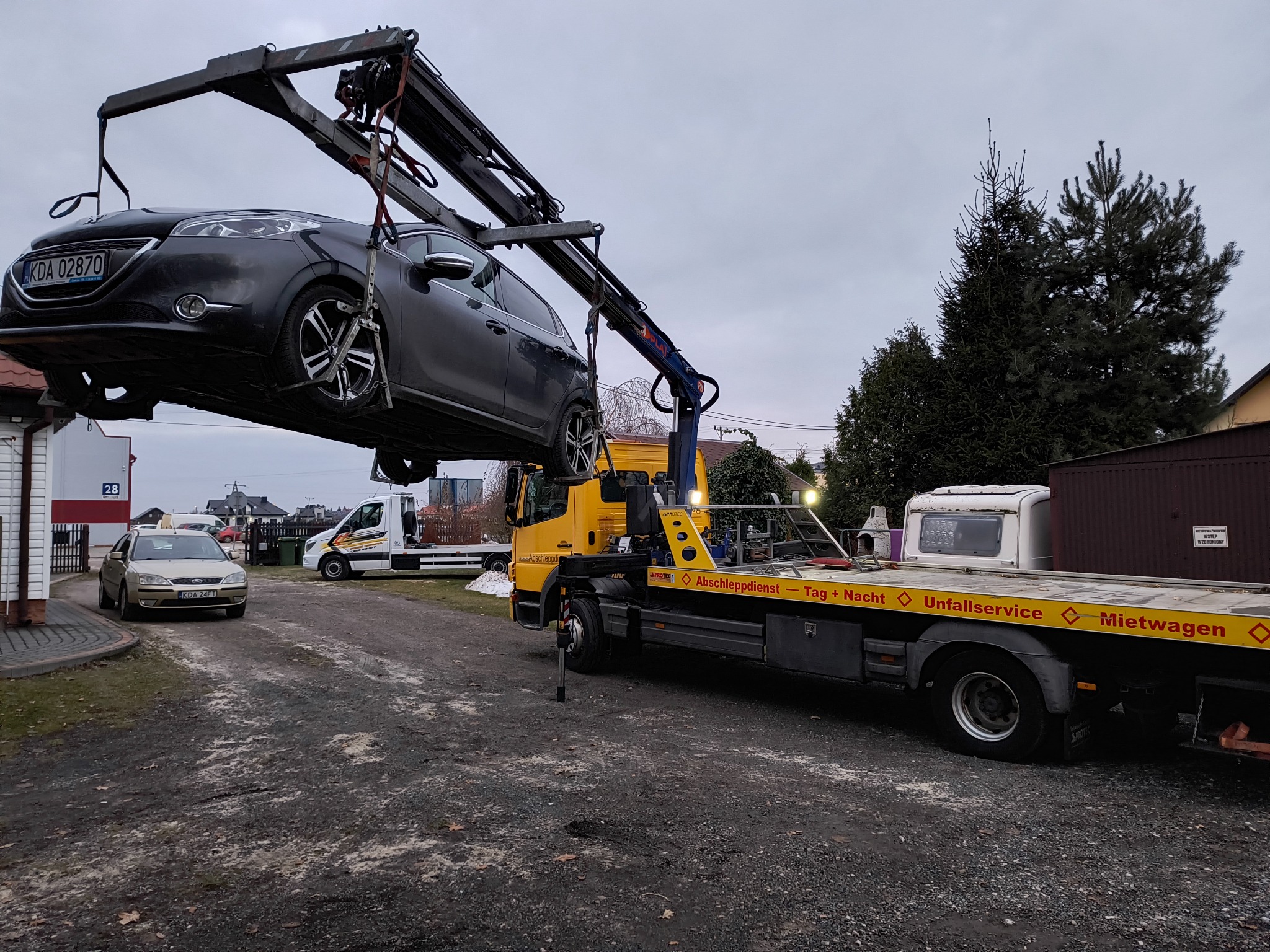 Szara osobówka podnoszona przez ramię żółtej lawety na tle zabudowań i drzew, widoczny numer rejestracyjny pojazdu oraz napisy na lawecie: Abschleppdienst, Unfallservice, Mietwagen.