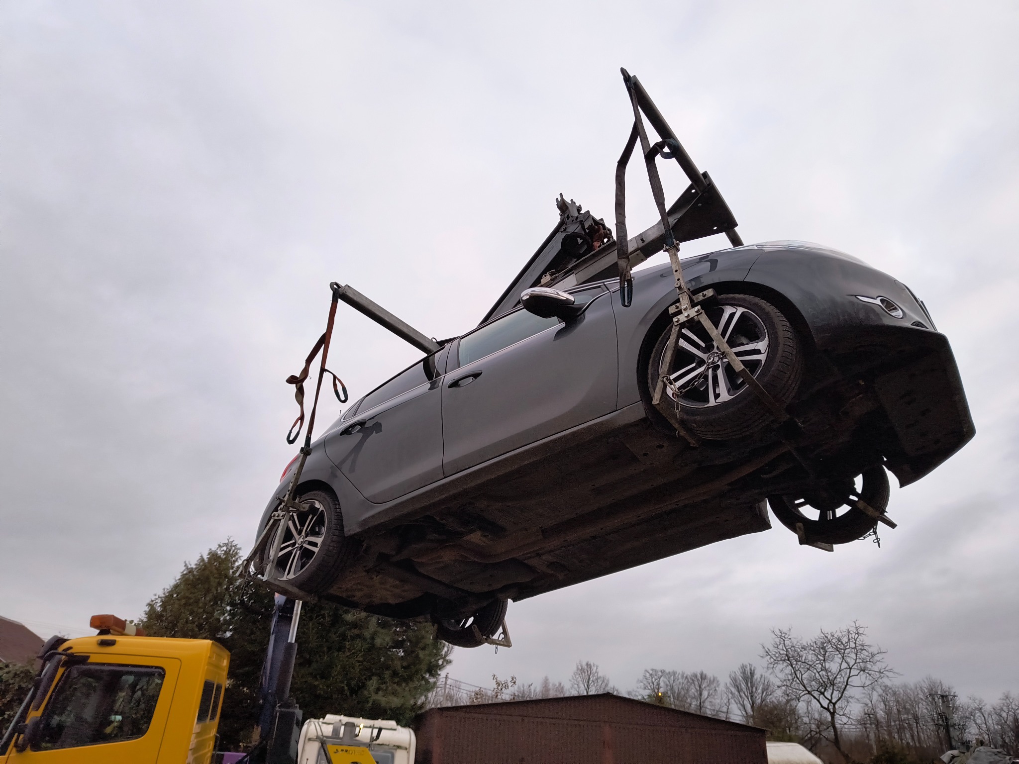 Szary samochód osobowy podnoszony przez żółtą lawetę na tle pochmurnego nieba i zabudowań.