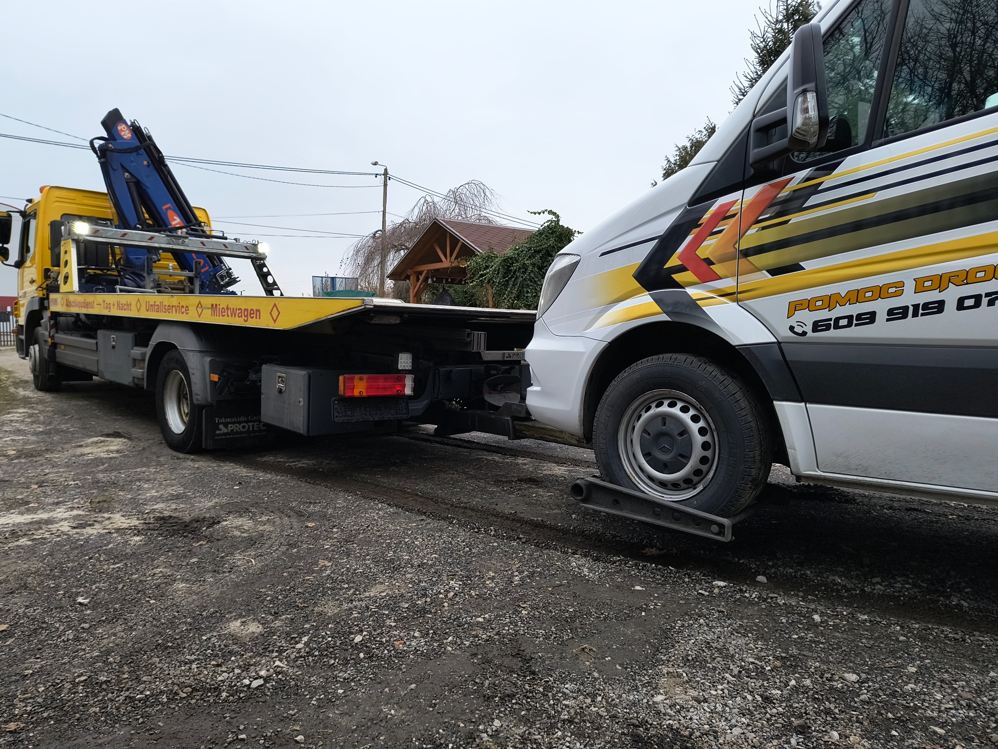 Żółta laweta z niebieskim dźwigiem holuje biały bus na żwirowym podłożu. Na busie widoczne logo firmy pomocy drogowej.