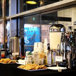Chefs Catering - Stół z czarnym obrusem, na którym znajdują się metalowe dystrybutory napojów (herbata, kawa), stosy papierowych kubków, ciasteczka na talerzykach i kartoniki z napojami roślinnymi, ustawione...