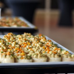 Chefs Catering - Taca z małymi kanapeczkami z pastą i posiekanymi warzywami (marchewka, ogórek, zioła) na białym prostokątnym talerzu, w tle rozmyte inne tace i elementy wyposażenia sali.