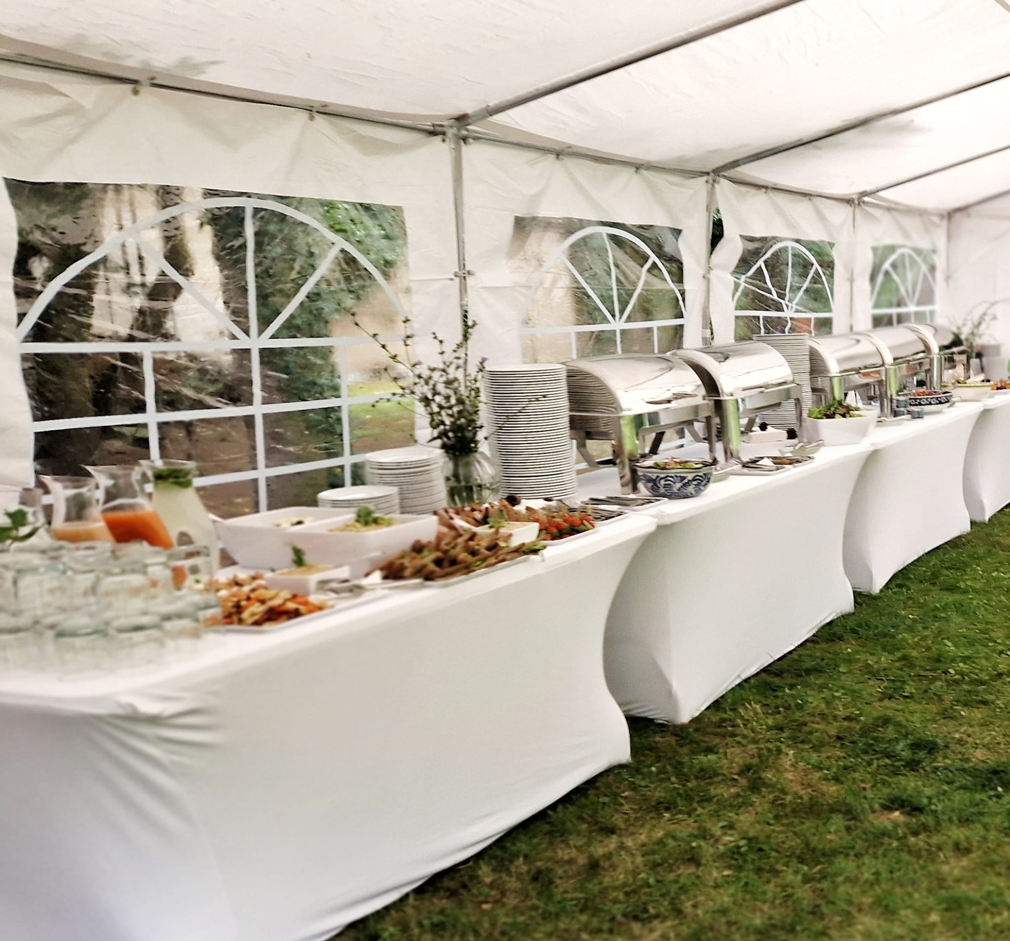 Długi stół bufetowy z białym obrusem wewnątrz namiotu, zastawiony różnorodnymi daniami, napojami w dzbankach, stosami talerzy i podgrzewaczami, widok z poziomu trawnika.