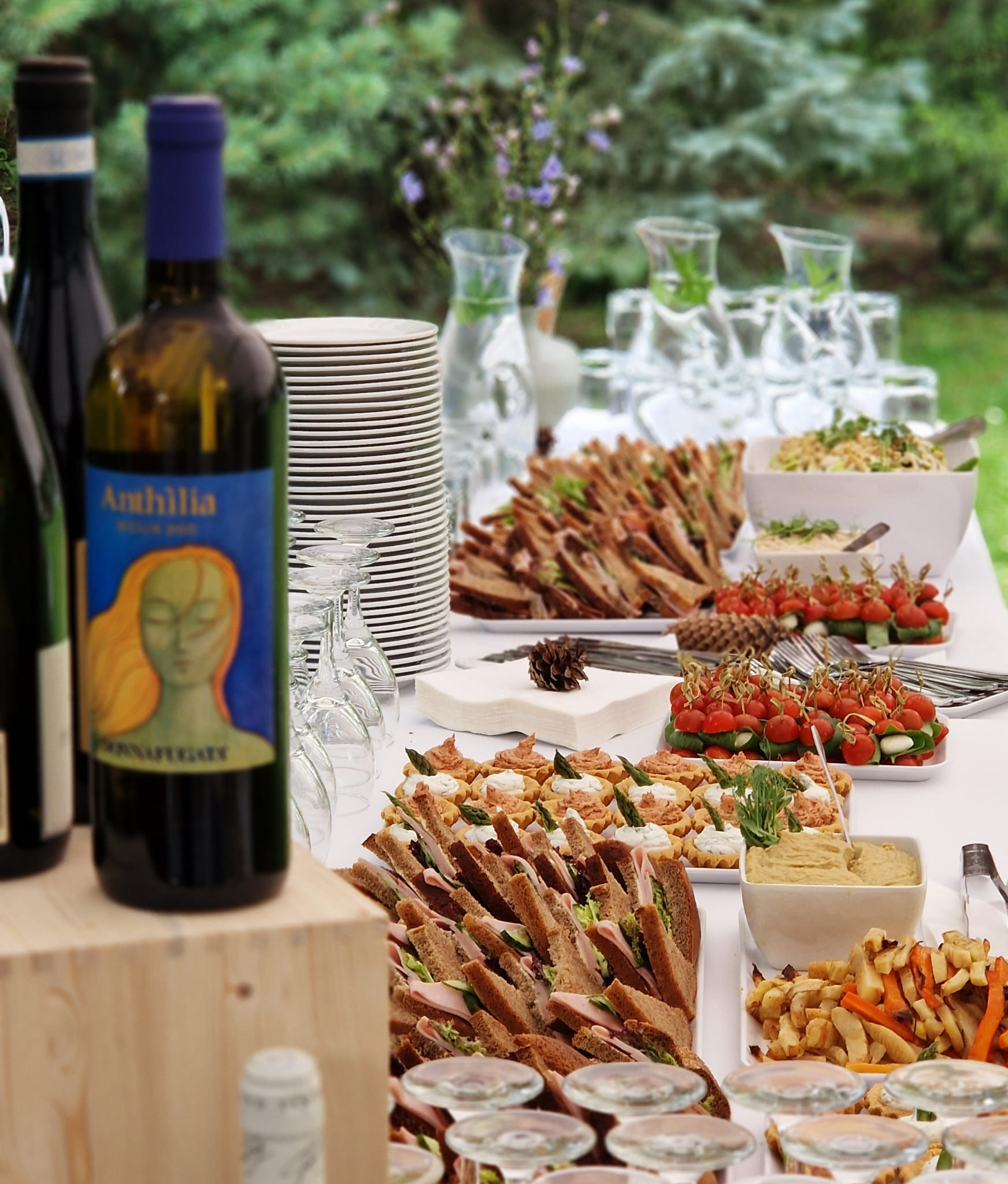 Stół cateringowy na zewnątrz: kanapki z ciemnego pieczywa, mini tartaletki z kremem i szparagami, koreczki z pomidorkami cherry i mozarellą, sałatka, frytki z warzyw korzeniowych, dzbanki z wodą...