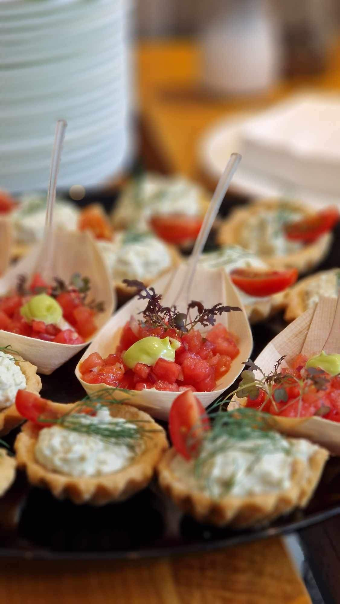 Bliski plan tacy z różnorodnymi, apetycznie wyglądającymi przekąskami: tartaletki z kremowym nadzieniem i koperkiem oraz łódeczki z drewna z pomidorami, awokado i ziołami, podane z plastikowymi...