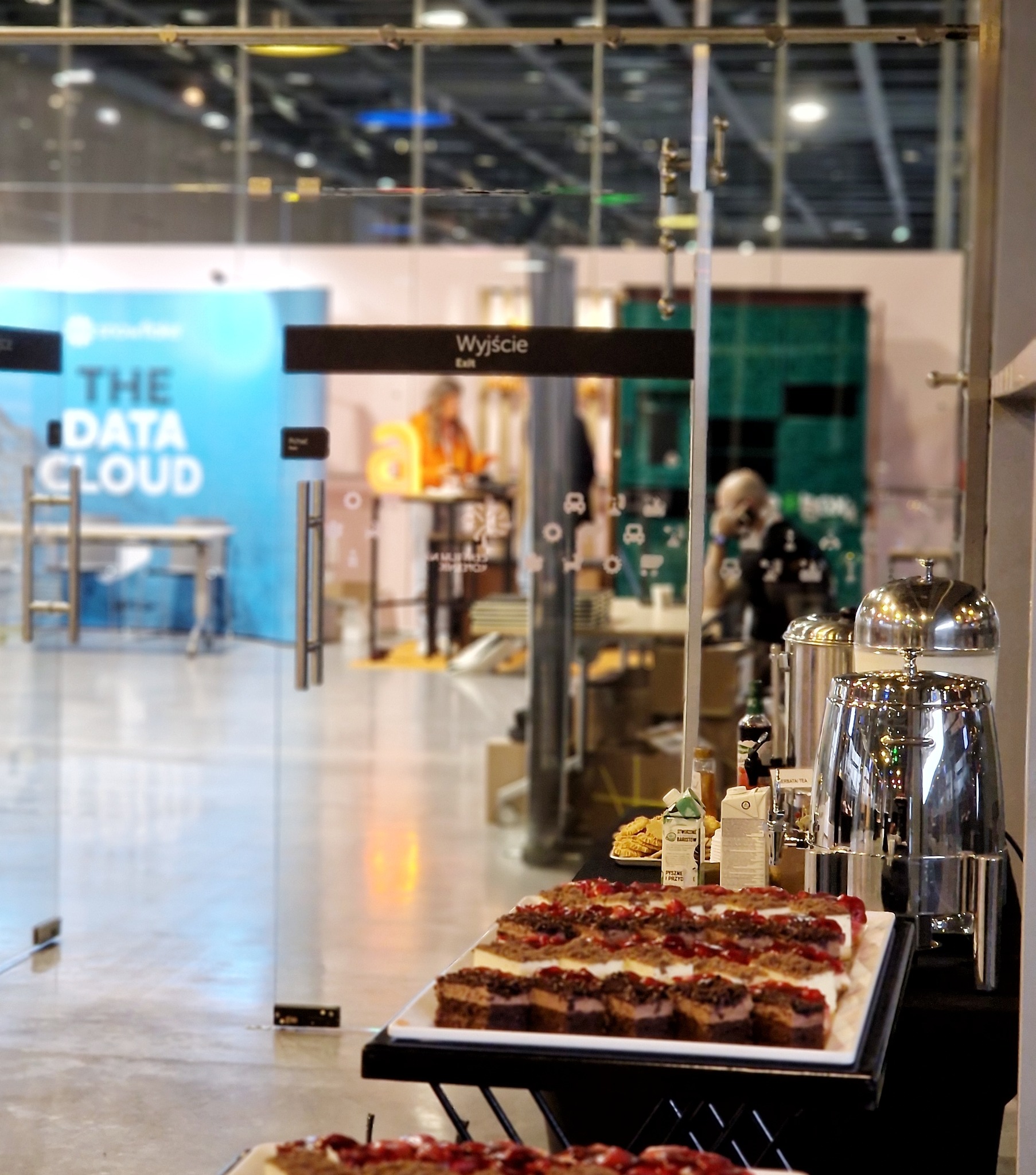 Stół cateringowy z ciastami i napojami na tle przeszklonego wejścia z napisem 'Wyjście' i logo 'The Data Cloud'.