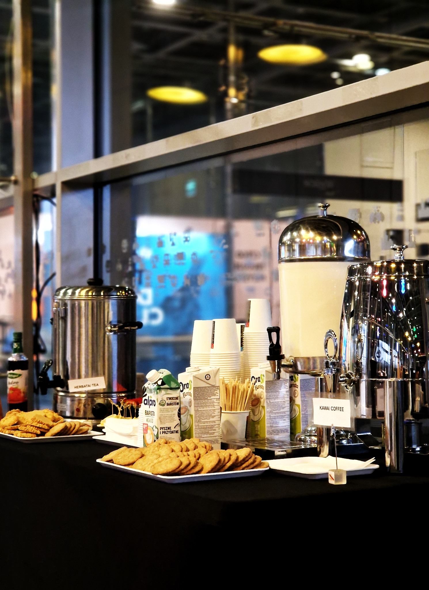 Stół z czarnym obrusem, na którym znajdują się metalowe dystrybutory napojów (herbata, kawa), stosy papierowych kubków, ciasteczka na talerzykach i kartoniki z napojami roślinnymi, ustawione...